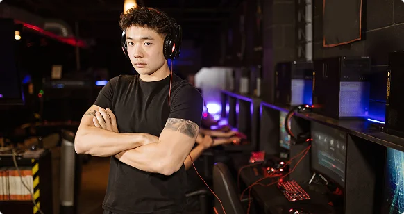 Un giovane giocatore di eSport asiatico con le cuffie indosso è in piedi a braccia conserte in un internet café